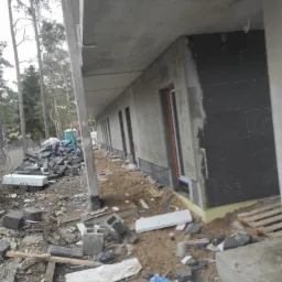 Budynek w trakcie ocieplania elewacji: widoczna czarna izolacja termiczna na ścianie, okno z pomarańczową ramą, stos kamieni i gruzu na ziemi, drzewa w tle.