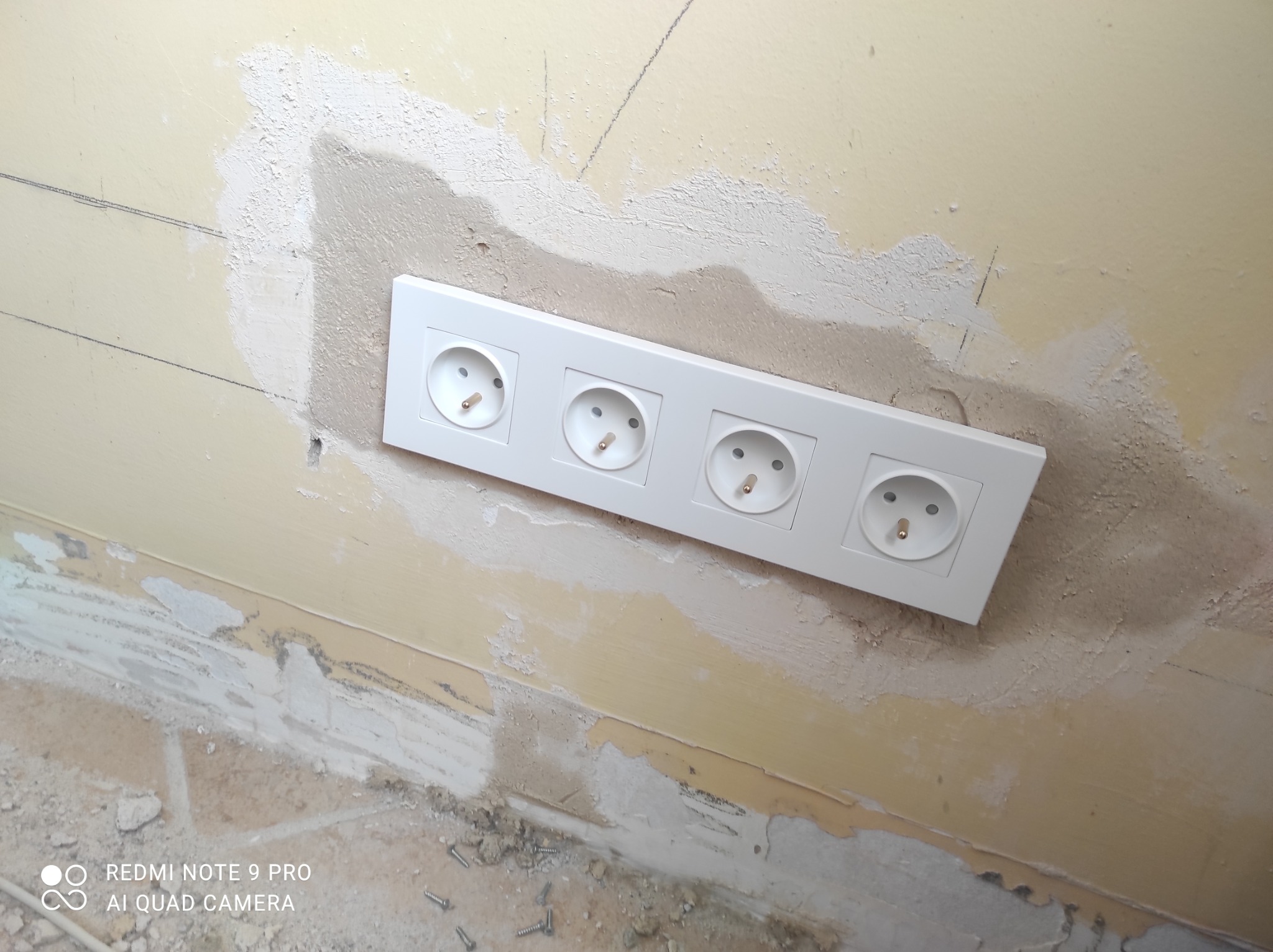 Blok czterech gniazdek elektrycznych zamontowany w ścianie, z widocznymi śladami po pracach wykończeniowych, szpachlą i niedomalowaną powierzchnią.