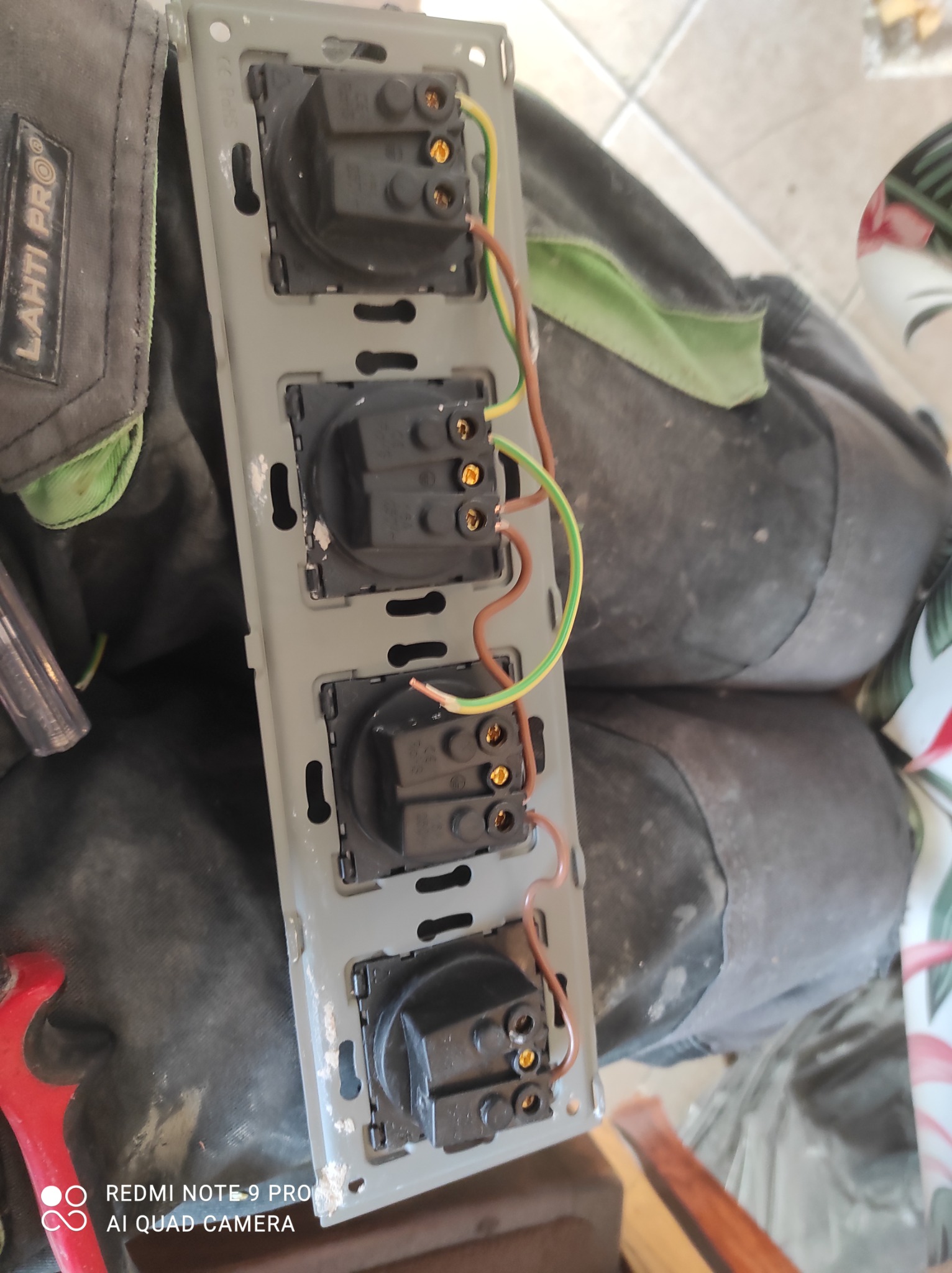 Potrójne gniazdko elektryczne w szarej ramce z podłączonymi przewodami, widoczne narzędzia i torba monterska Lahti Pro w tle.