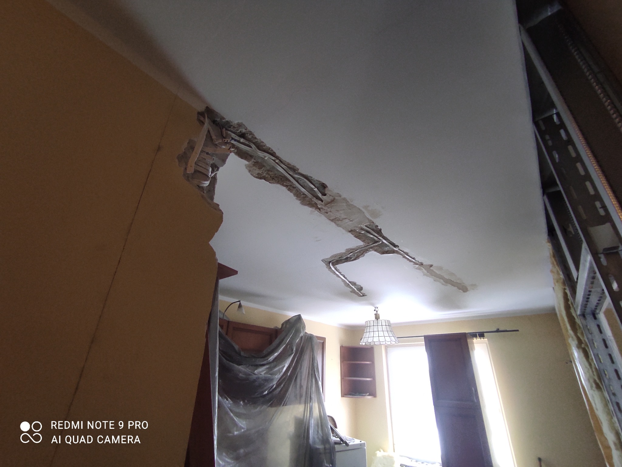 Uszkodzony sufit z odsłoniętymi przewodami i fragmentami konstrukcji, zabezpieczone meble w tle, widoczne ślady demontażu.