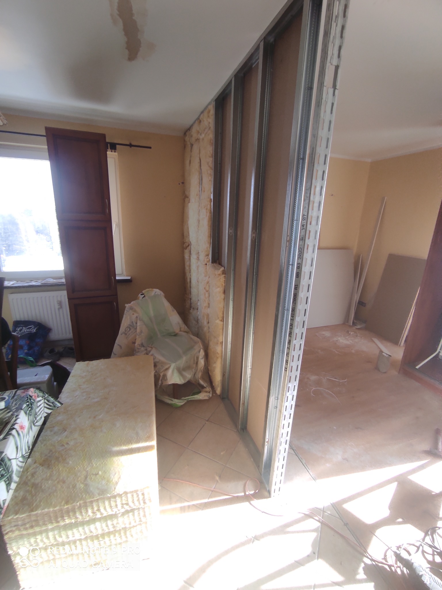 Wnętrze w trakcie tworzenia ścianki działowej z profili metalowych i płyt kartonowo-gipsowych, widoczne ocieplenie w ścianie, materiały budowlane i narzędzia w pomieszczeniu z brązowymi plamami...