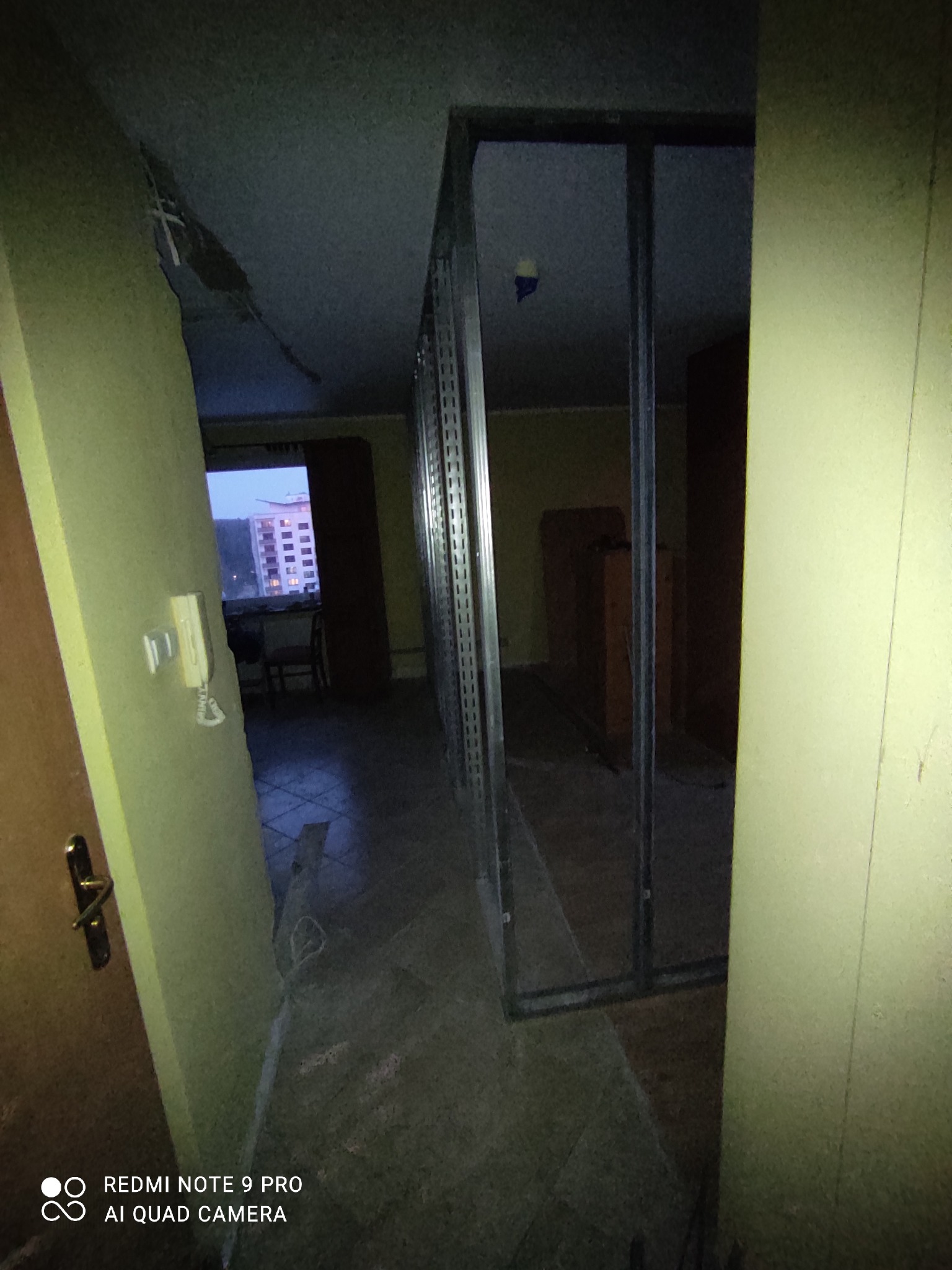 Wnętrze mieszkania w trakcie prac: widoczne metalowe profile konstrukcyjne ścianki działowej, surowy stan wykończenia, drzwi wejściowe z domofonem po lewej, w tle okno z widokiem na budynek...