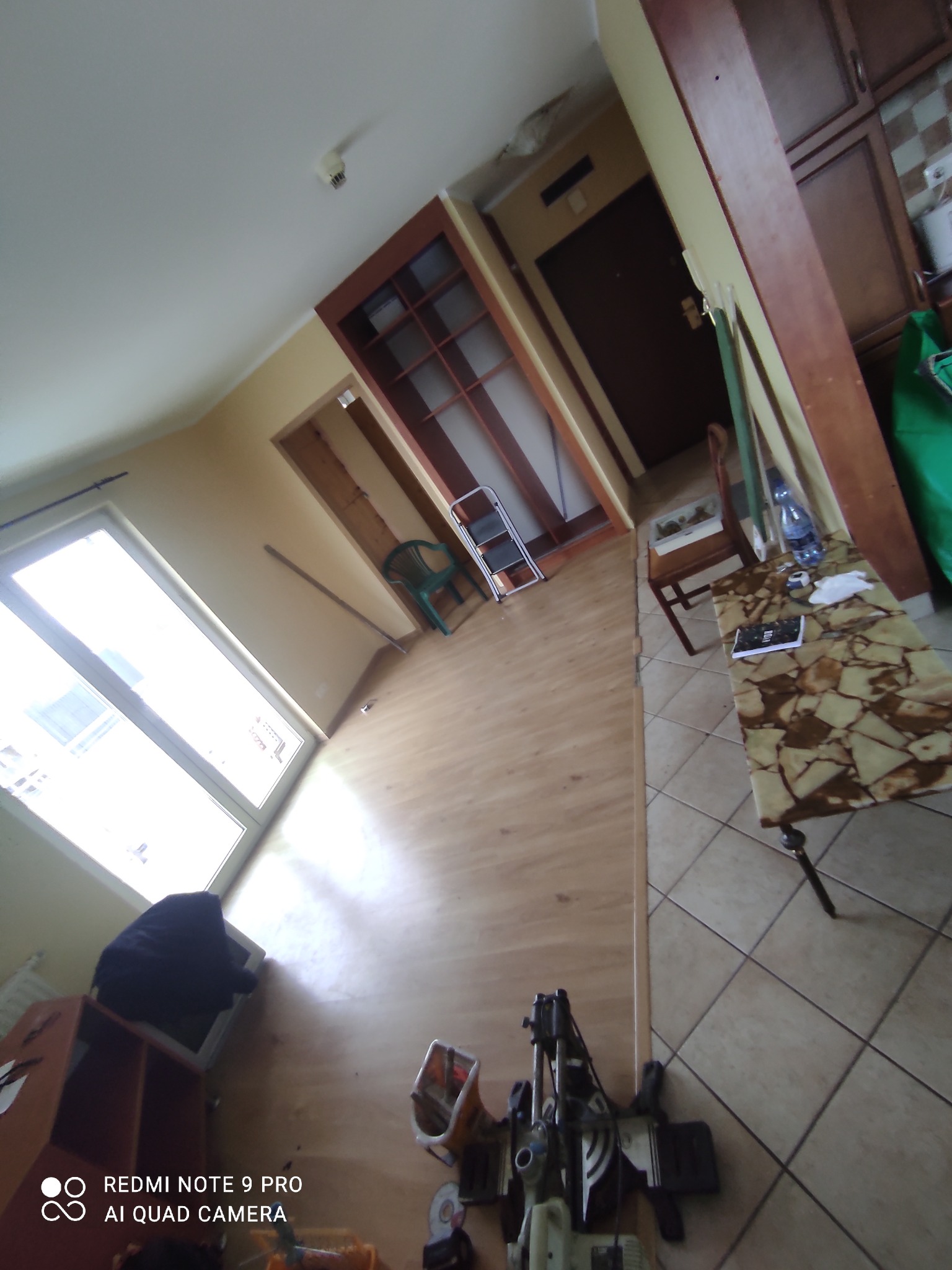 Wnętrze w trakcie prac wykończeniowych: widoczna podłoga, okno, narzędzia i materiały budowlane, częściowo umeblowane pomieszczenie z widocznym sprzętem do cięcia paneli.