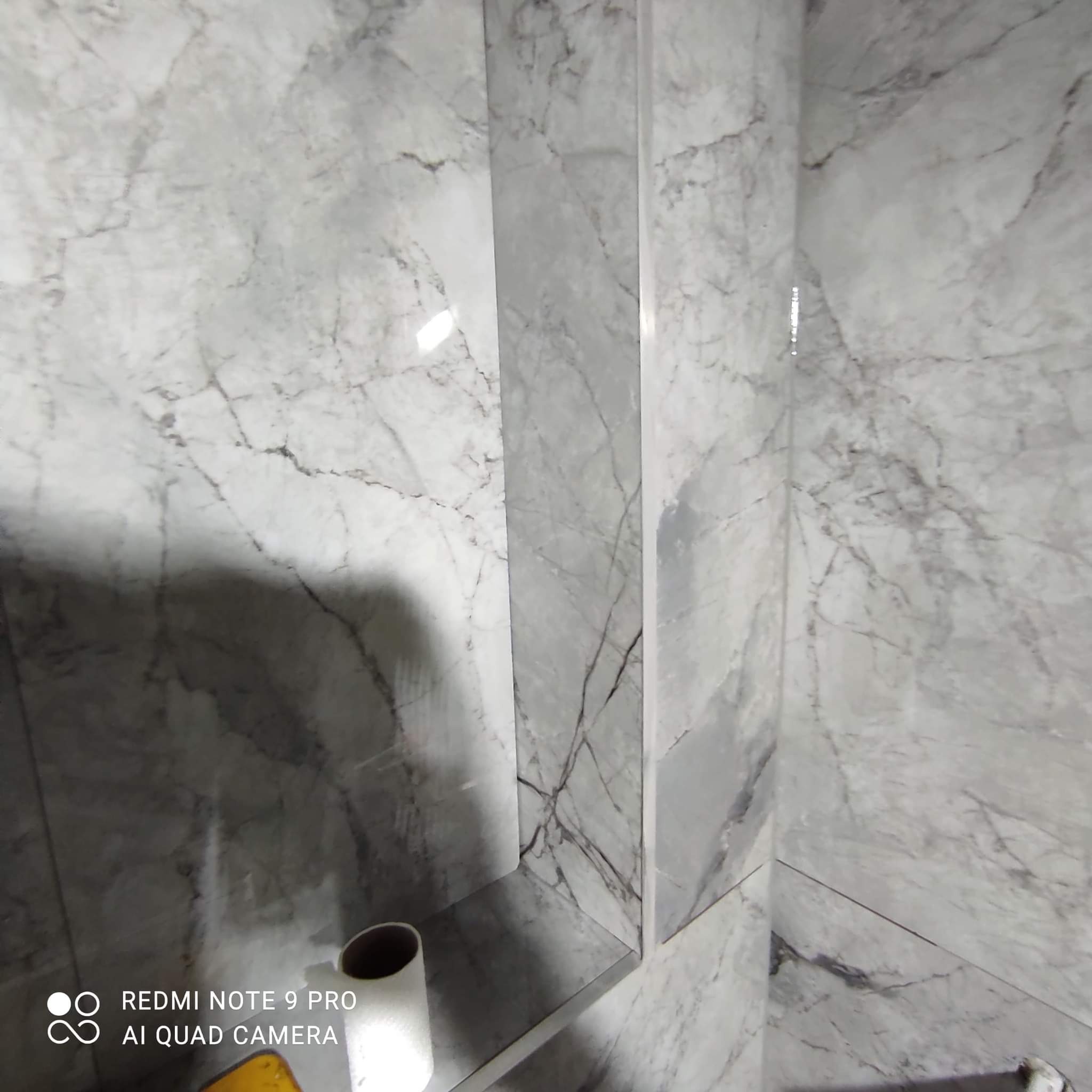 Wykończona ściana łazienki z szarymi płytkami imitującymi marmur, z wbudowaną półką i widoczną rolką papieru toaletowego.