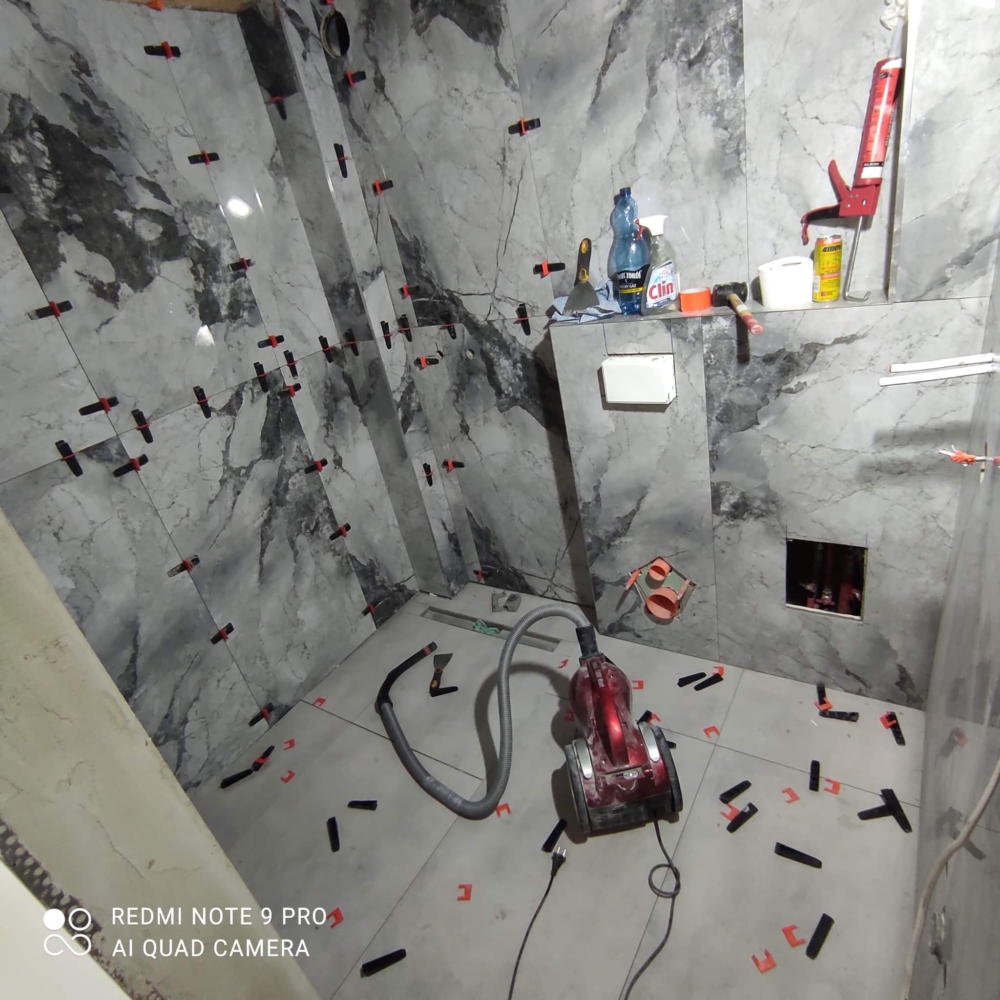 Łazienka w trakcie układania płytek imitujących marmur, widoczne krzyżyki dystansowe, poziomica, odkurzacz i akcesoria glazurnicze na półce.