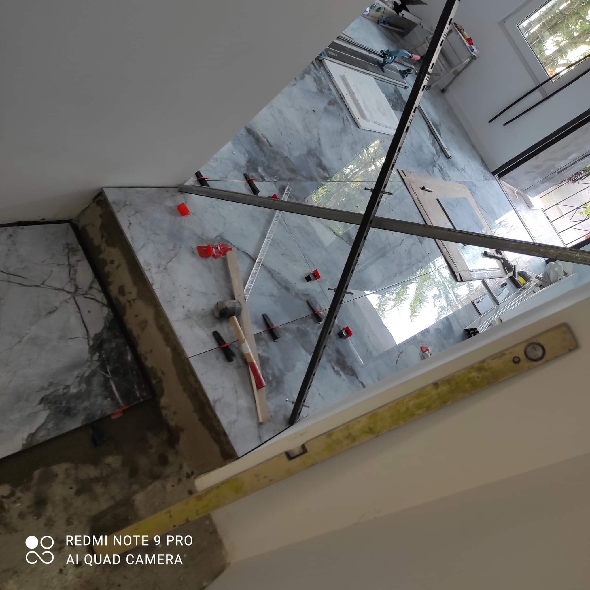 Układanie płytek imitujących marmur na podłodze, widoczne narzędzia glazurnicze, poziomica, młotek gumowy, krzyżyki dystansowe, listwy, częściowo ułożona podłoga, okno w tle.