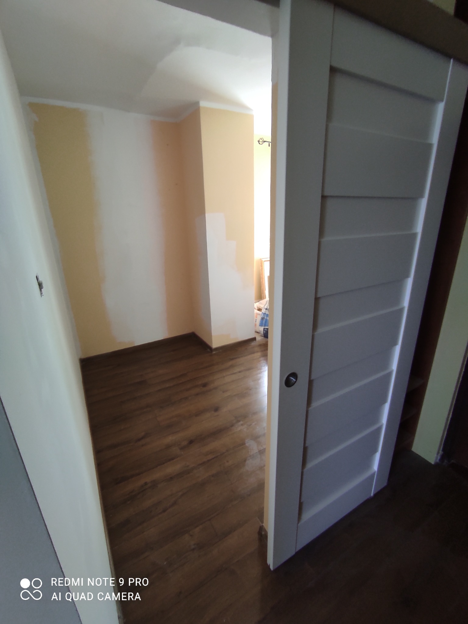 Wnętrze pokoju z drewnianą podłogą i częściowo pomalowanymi ścianami, widoczne przez otwarte białe drzwi przesuwne z poziomymi panelami.