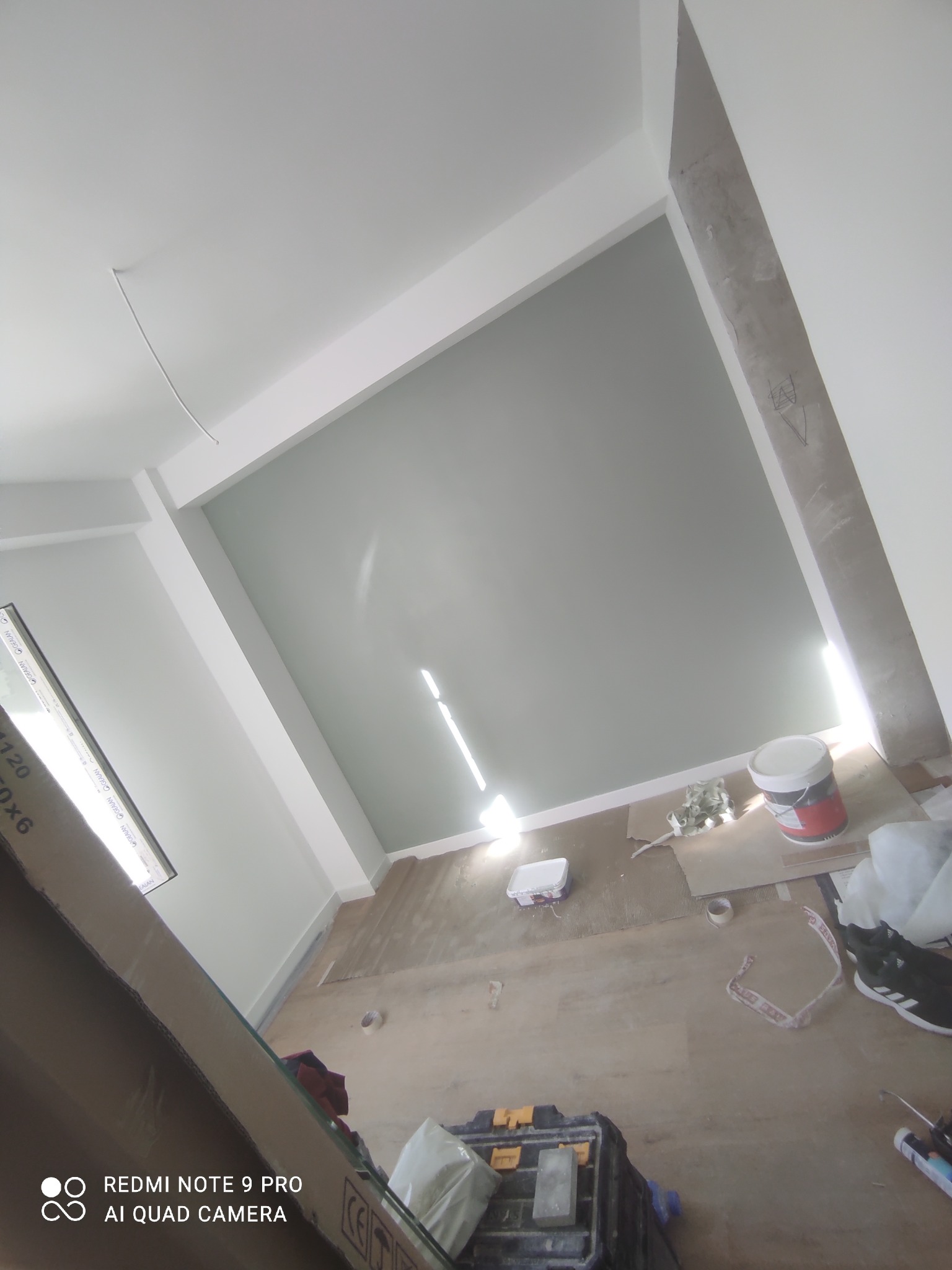 Wnętrze w trakcie odświeżania: pomalowana na szaro ściana z białym pasem sufitowym, widoczna puszka z farbą, narzędzia i resztki materiałów na podłodze.