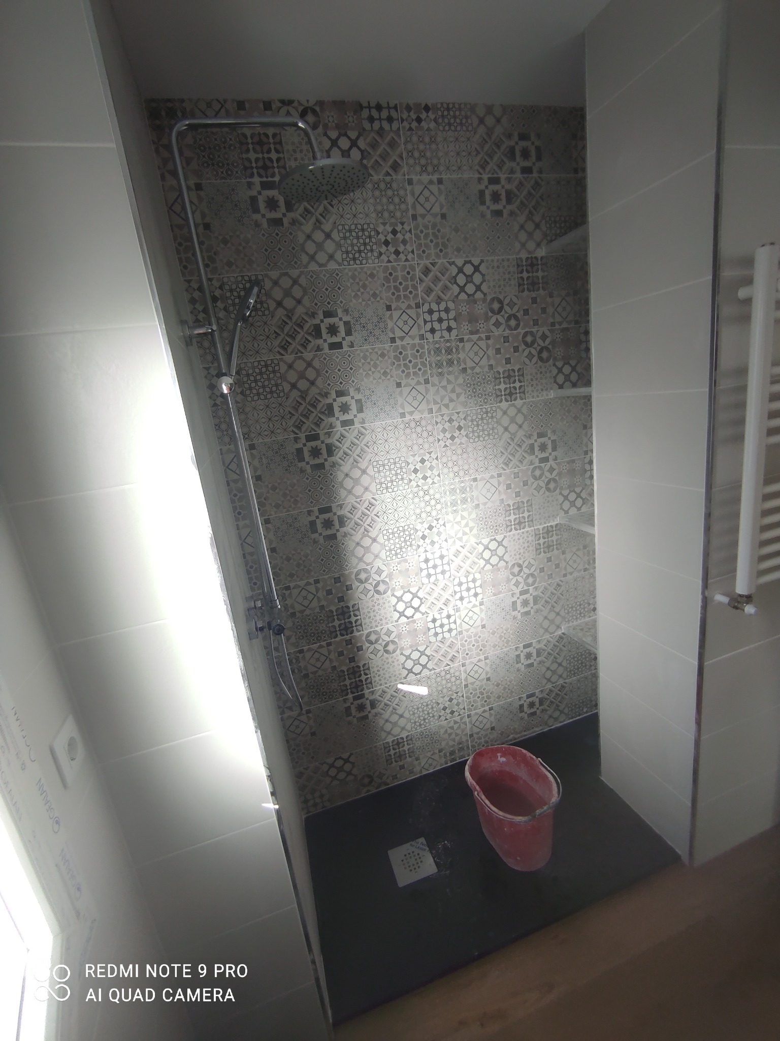 Wykończony prysznic w łazience z wzorzystymi płytkami na ścianie, ciemną podłogą i metalowym zestawem prysznicowym; czerwone wiadro stoi na podłodze.