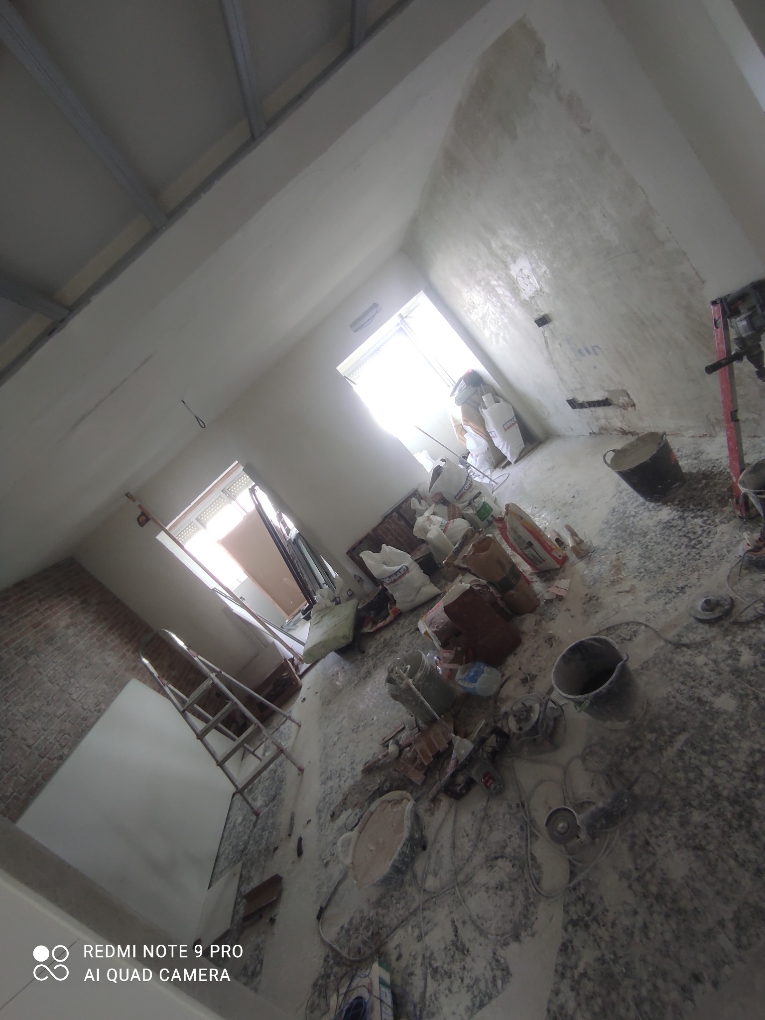 Wnętrze w trakcie prac wykończeniowych: surowe ściany, worki z materiałami budowlanymi, narzędzia i drabina, widoczne profile sufitowe podwieszane oraz pracownik w oddali przy oknie.