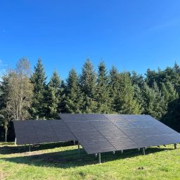 G-PRO ECO ENERGY - Dwa rzędy paneli słonecznych na metalowych stelażach, ustawione na trawiastym terenie z drzewami w tle, pod błękitnym niebem.