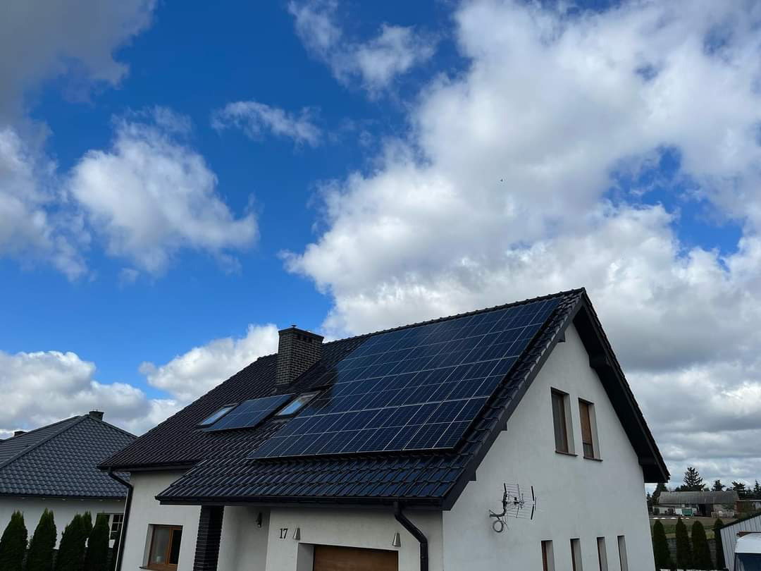 Dom jednorodzinny z panelami fotowoltaicznymi na dachu, widoczne okna dachowe, antena telewizyjna na ścianie, błękitne niebo z chmurami w tle.