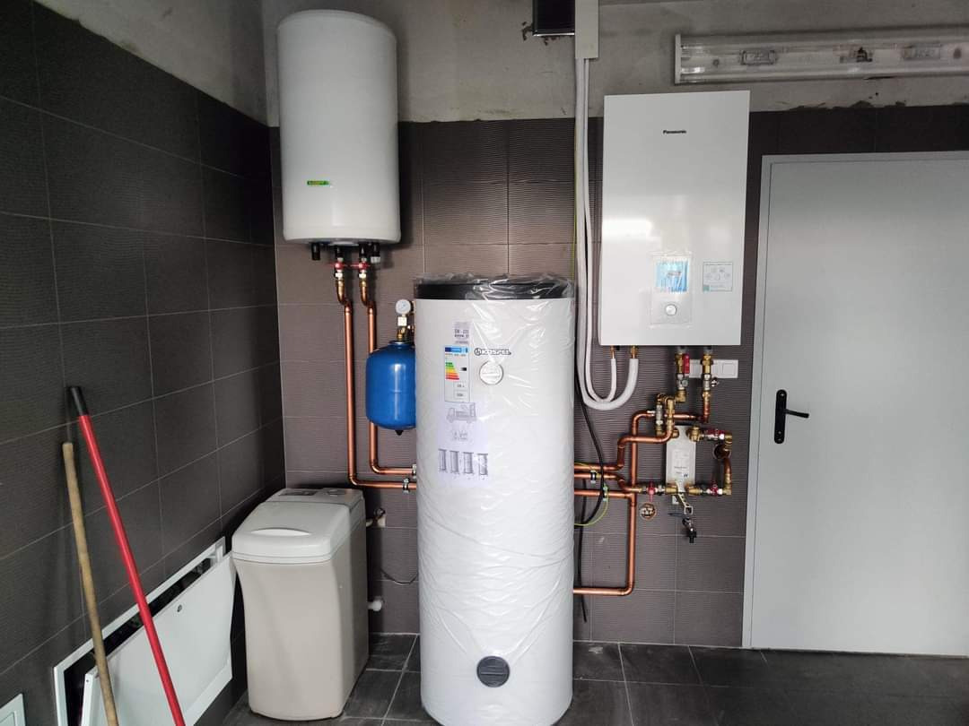 Kompleksowa instalacja grzewcza w kotłowni w Kaliszu: dwa zbiorniki, piec gazowy Panasonic, zmiękczacz wody i liczne miedziane rury łączące urządzenia.
