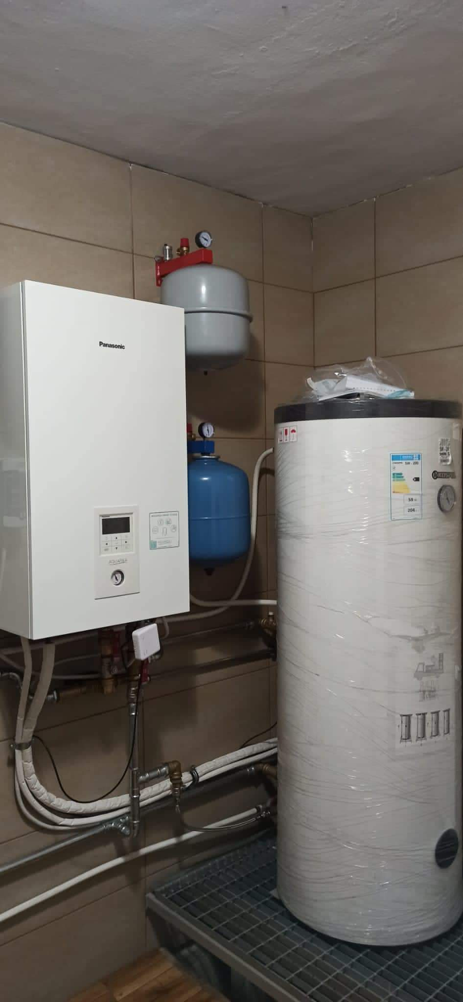 Biały piec gazowy Panasonic, szary zbiornik wyrównawczy, niebieski zbiornik ciśnieniowy oraz biały, owinięty folią zbiornik ciepłej wody na metalowym podeście w kotłowni wyłożonej kafelkami.