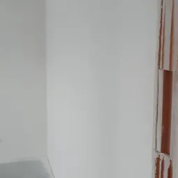 Świeżo pomalowana biała ściana w rogu pomieszczenia, widoczny fragment sufitu i podłogi, surowy mur z cegieł po prawej stronie.