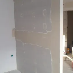 Ściana z płyty gipsowo-kartonowej w trakcie szpachlowania, widoczne miejsca łączeń i śrub, kabel elektryczny zwisający z sufitu, fragment podłogi z resztkami gipsu.