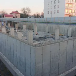 Betonowy fundament budynku w trakcie budowy, widoczne zbrojenia, słupy i izolacja pionowa, w tle budynki mieszkalne i pracownicy w pomarańczowych kombinezonach.