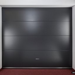OknaGK - OKNOPLAST Góra Kalwaria - Zamknięta, segmentowa brama garażowa w kolorze grafitowym z subtelnymi poziomymi przetłoczeniami, widoczna z przodu na tle białych ścian i czerwonej wykładziny podłogowej.