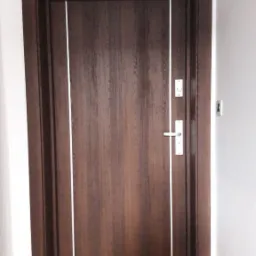 Ciemnobrązowe drzwi wewnętrzne z białymi intarsjami i srebrną klamką na tle białej ściany i drewnianej podłogi, z wycieraczką pod spodem.