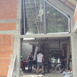 OknaGK - OKNOPLAST Góra Kalwaria - Montaż okna o nietypowym trójkątnym kształcie w surowym stanie budynku, widoczne ekipy montażowe i opakowania produktów FAKRO.