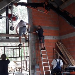 OknaGK - OKNOPLAST Góra Kalwaria - Montaż okna z użyciem specjalistycznego dźwigu przyssawkowego w surowym stanie budynku z cegły, pracownicy na rusztowaniach i drabinach, widoczne deski i materiały budowlane.