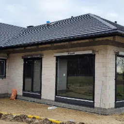 Surowy stan elewacji domu jednorodzinnego z zamontowanymi oknami PCV z czarnymi ramami i szarymi roletami zewnętrznymi, widoczne worki budowlane i żółte rury na podłożu.