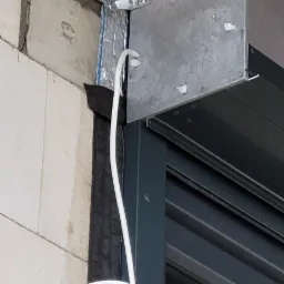 Fragment montażu rolety zewnętrznej z widoczną puszką i przewodem elektrycznym, zamocowaną do ściany z betonu komórkowego.