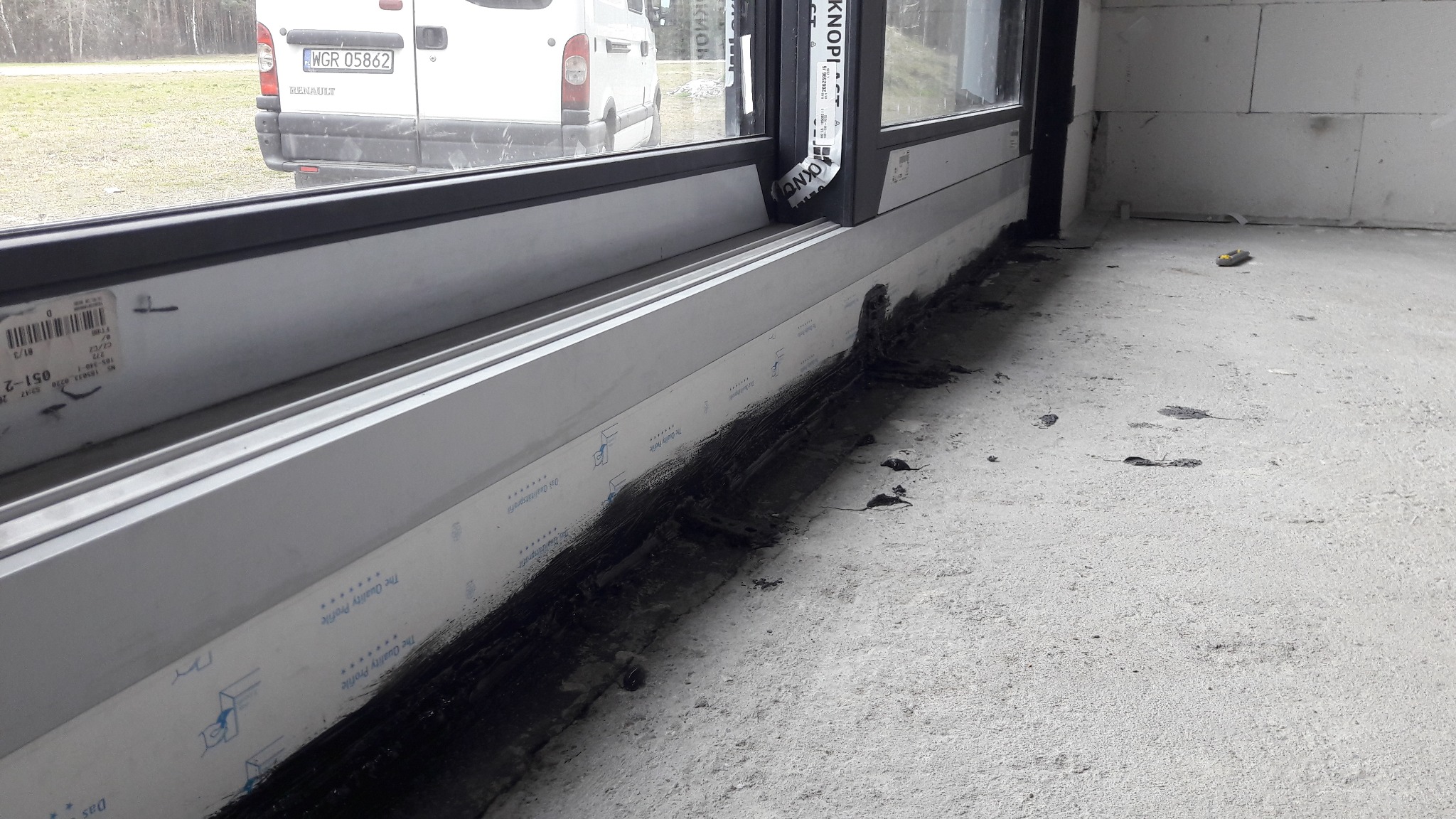 Montaż okna w surowym stanie w Górze Kalwarii, widoczne uszczelnienie pianką montażową i folią, na zewnątrz zaparkowany biały samochód dostawczy.