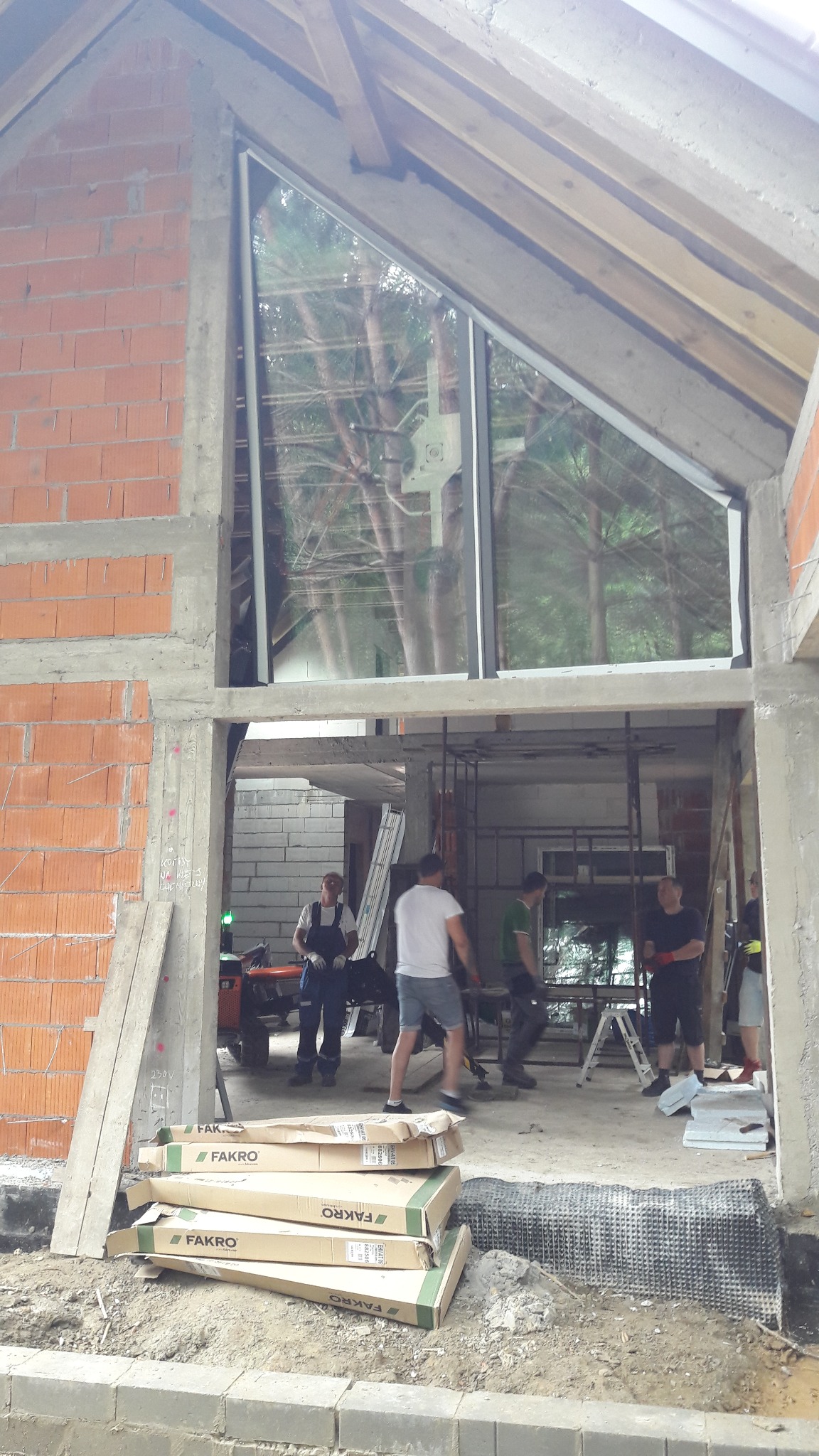 Montaż okna o nietypowym trójkątnym kształcie w surowym stanie budynku, widoczne ekipy montażowe i opakowania produktów FAKRO.