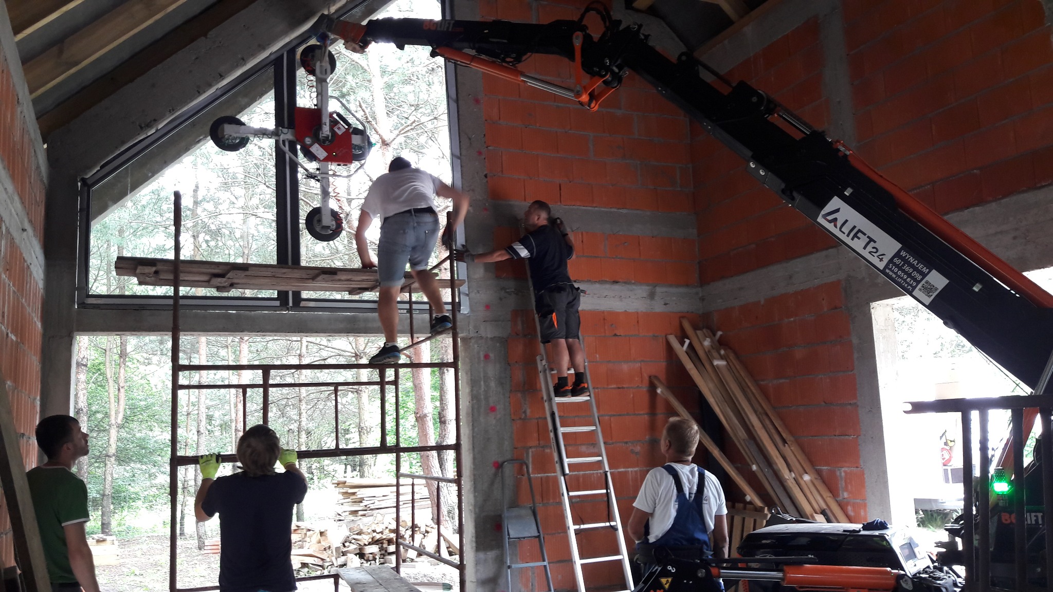 Montaż okna z użyciem specjalistycznego dźwigu przyssawkowego w surowym stanie budynku z cegły, pracownicy na rusztowaniach i drabinach, widoczne deski i materiały budowlane.