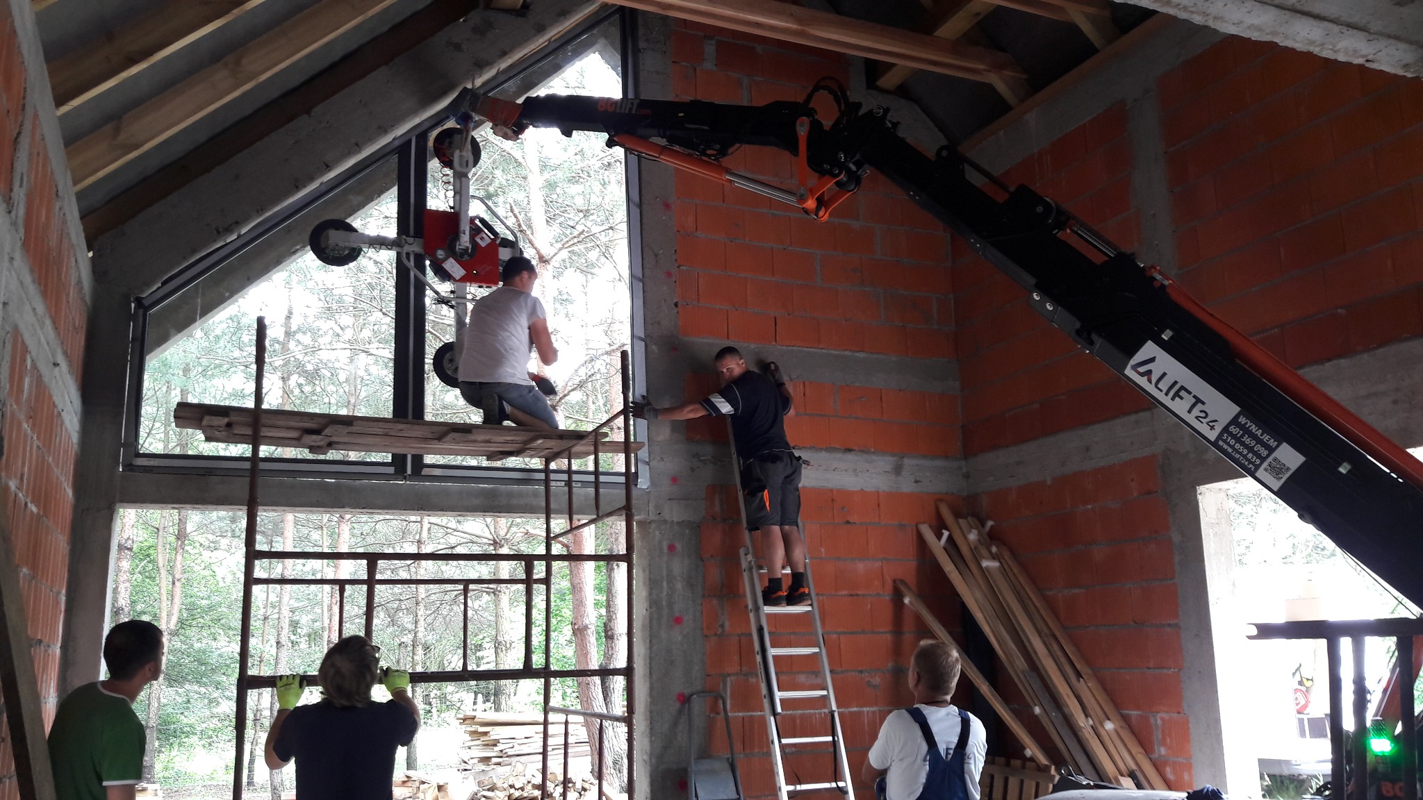 Montaż okna w surowym stanie budynku z wykorzystaniem podnośnika próżniowego i rusztowania, widoczni pracownicy w trakcie pracy.