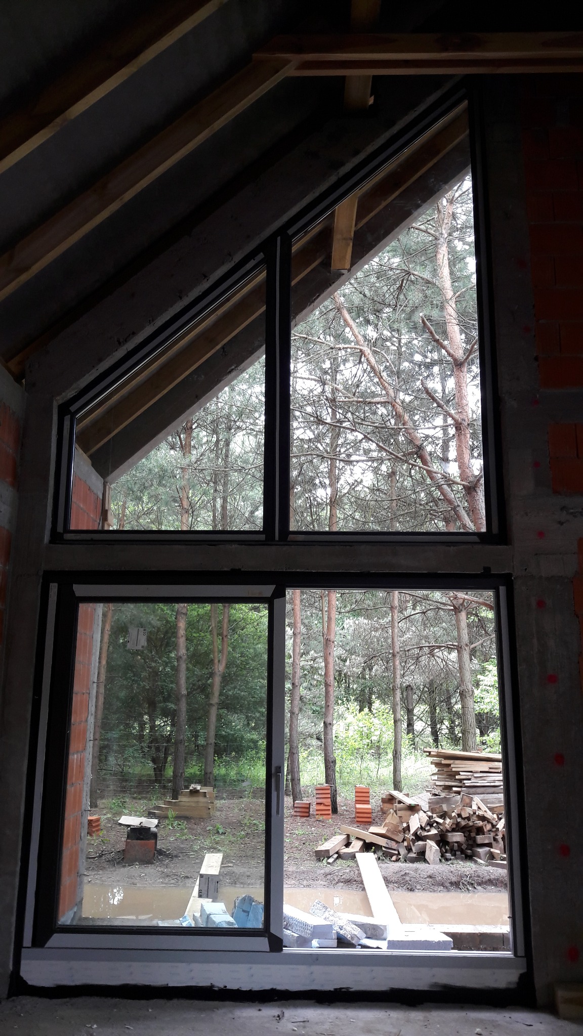 Czarna rama okienna z widokiem na las, w trakcie montażu w surowym budynku z cegły i betonu, widoczne drewniane elementy konstrukcji dachu.