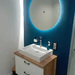 Nowoczesna łazienka z umywalką nablatową, szafką z drewnianym blatem i okrągłym lustrem z podświetleniem LED na tle ściany w kolorze głębokiego granatu.