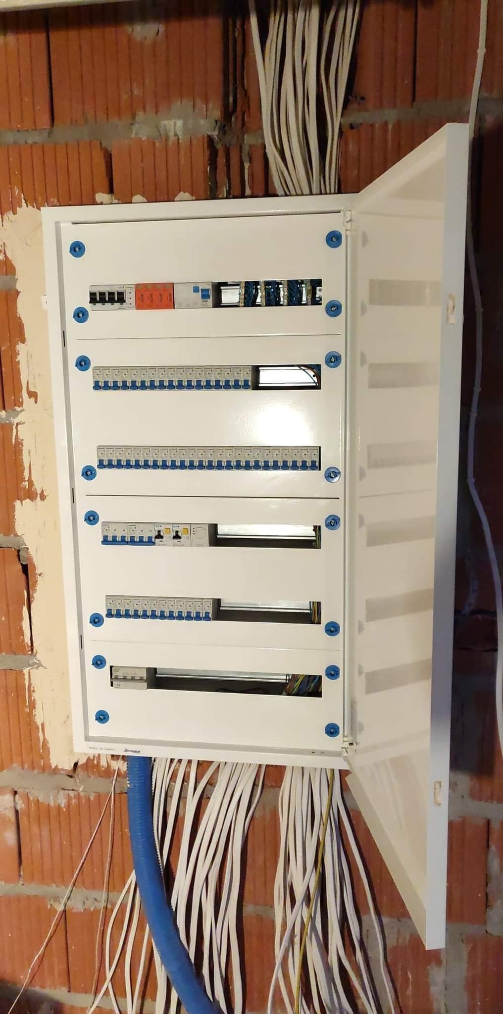 Otwarta, biała skrzynka elektryczna z bezpiecznikami zamontowana na ścianie z czerwonej cegły, z widocznymi kablami wchodzącymi do skrzynki od góry i od dołu, oraz niebieską rurą ochronną. Widoczne...