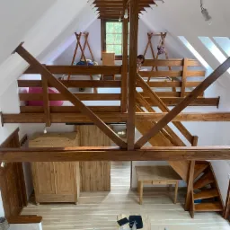 Wnętrze domu z antresolą i drewnianą konstrukcją nośną, widok z góry na stół z laptopem i krzesłami, oraz schody z drewna.