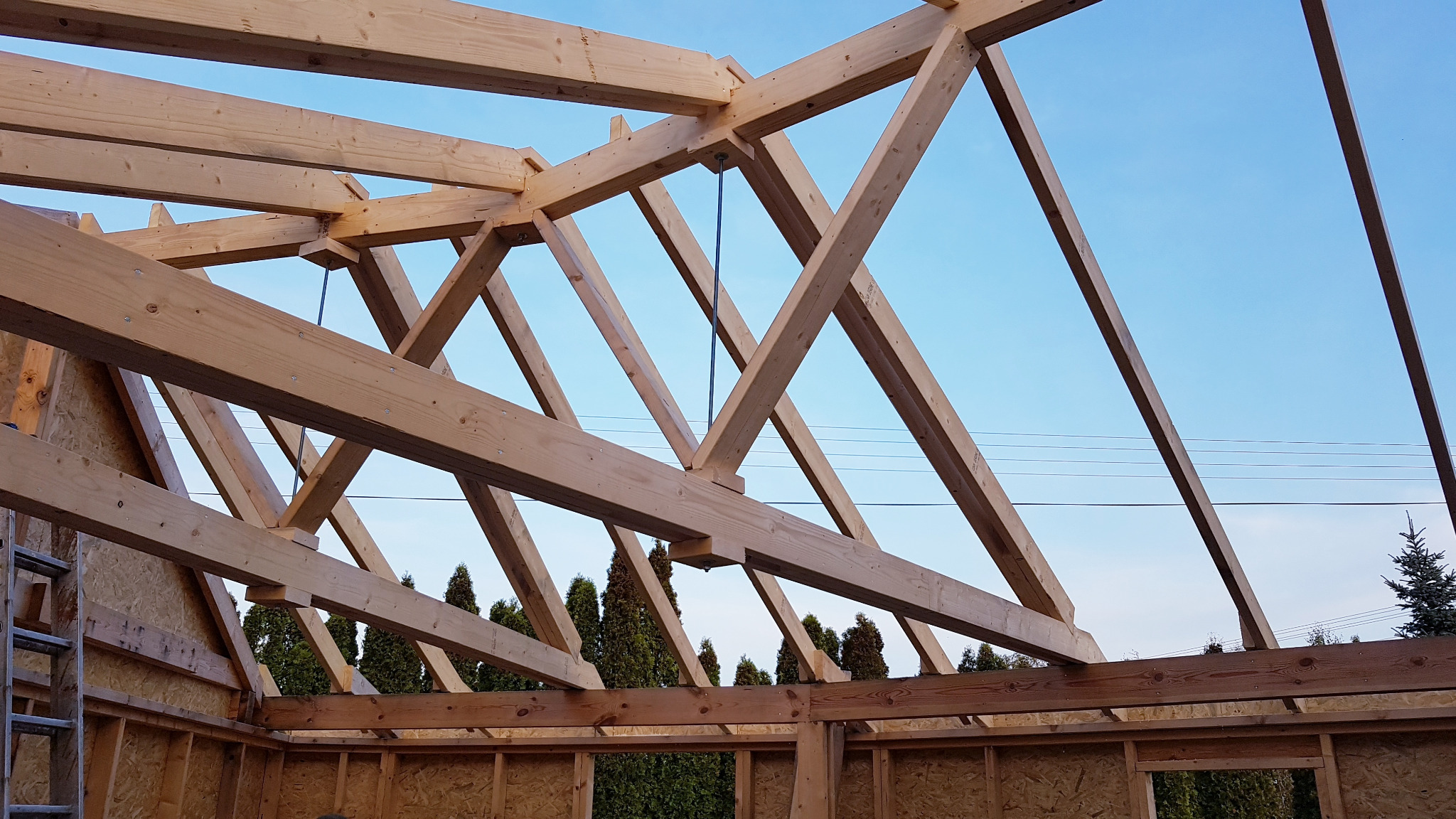 Konstrukcja dachu z drewna w trakcie budowy, widoczne elementy więźby dachowej, poszycie ścian płytami OSB, oraz fragment drabiny w rogu kadru, na tle błękitnego nieba i zielonych drzew.