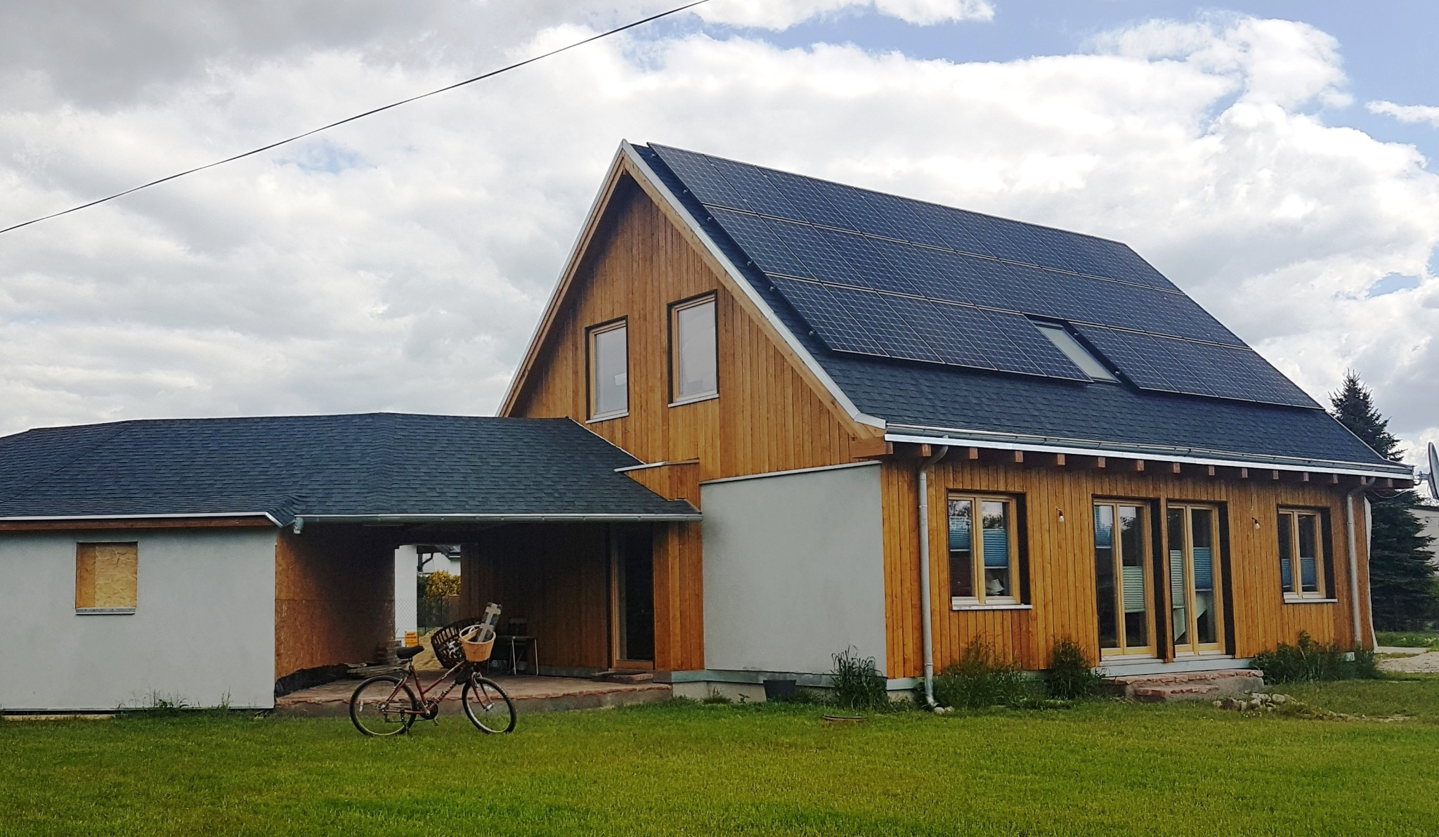 Nowoczesny dom z drewnianą elewacją i panelami słonecznymi na dachu, z rowerem opartym o ścianę garażu.