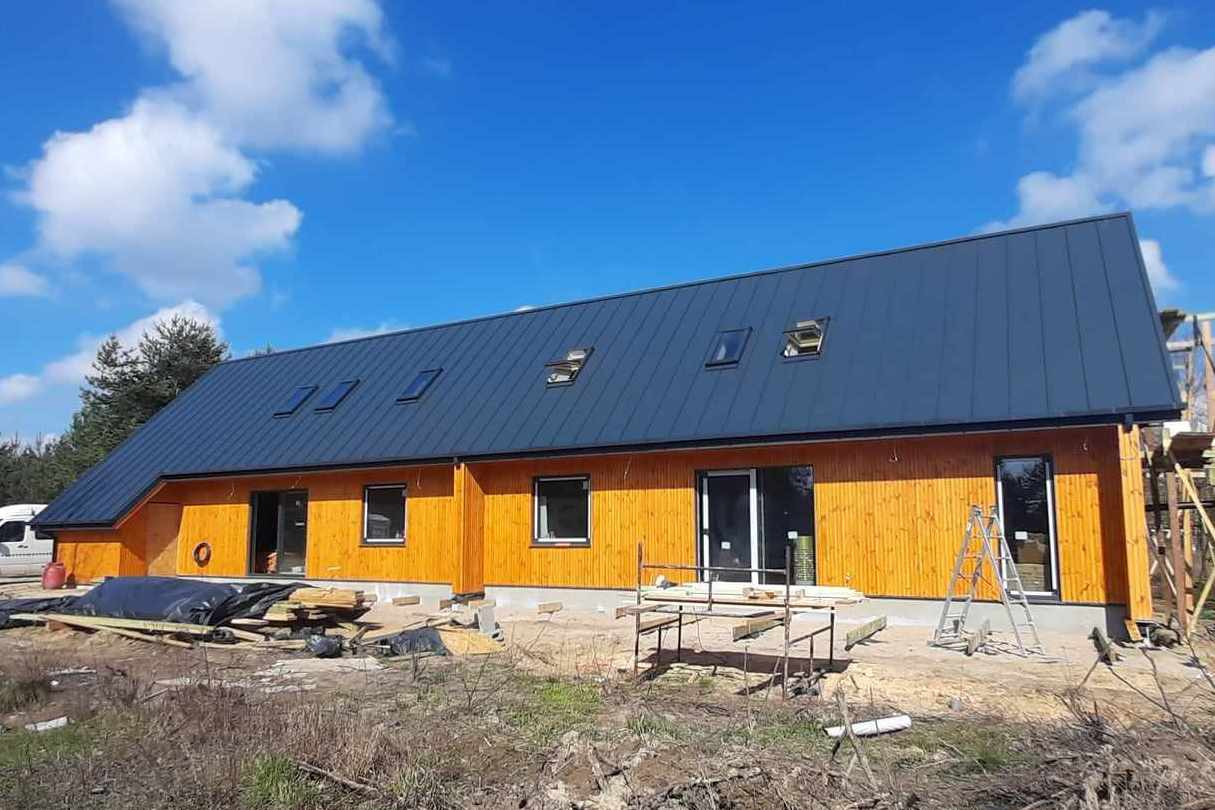 Surowy dom szkieletowy z drewnianą elewacją i ciemnym dachem z lukarnami, widoczne materiały budowlane i drabina przy ścianie.