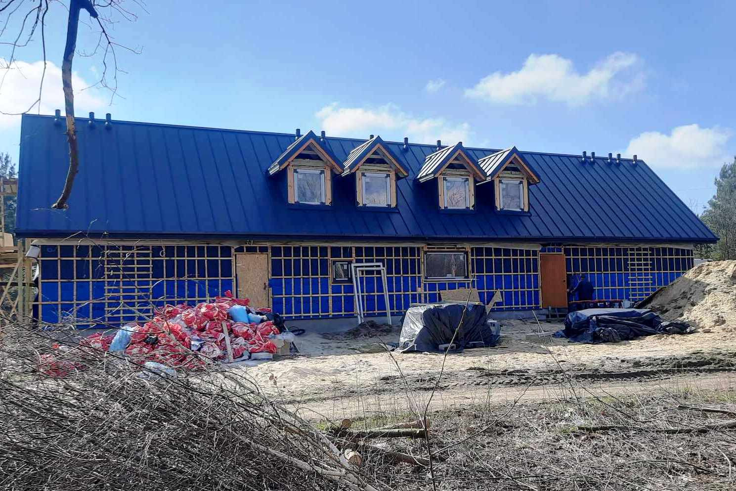 Budowa domu w trakcie realizacji: widoczna niebieska membrana na ścianach, drewniana konstrukcja szkieletowa, świeżo położony ciemnoniebieski dach z trzema lukarnami, sterta materiałów budowlanych...
