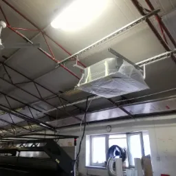 Centrala wentylacyjna w folii ochronnej, zawieszona na metalowych profilach pod sufitem hali przemysłowej. Widoczne elementy instalacji, okno z wentylatorem i kalendarz na ścianie.