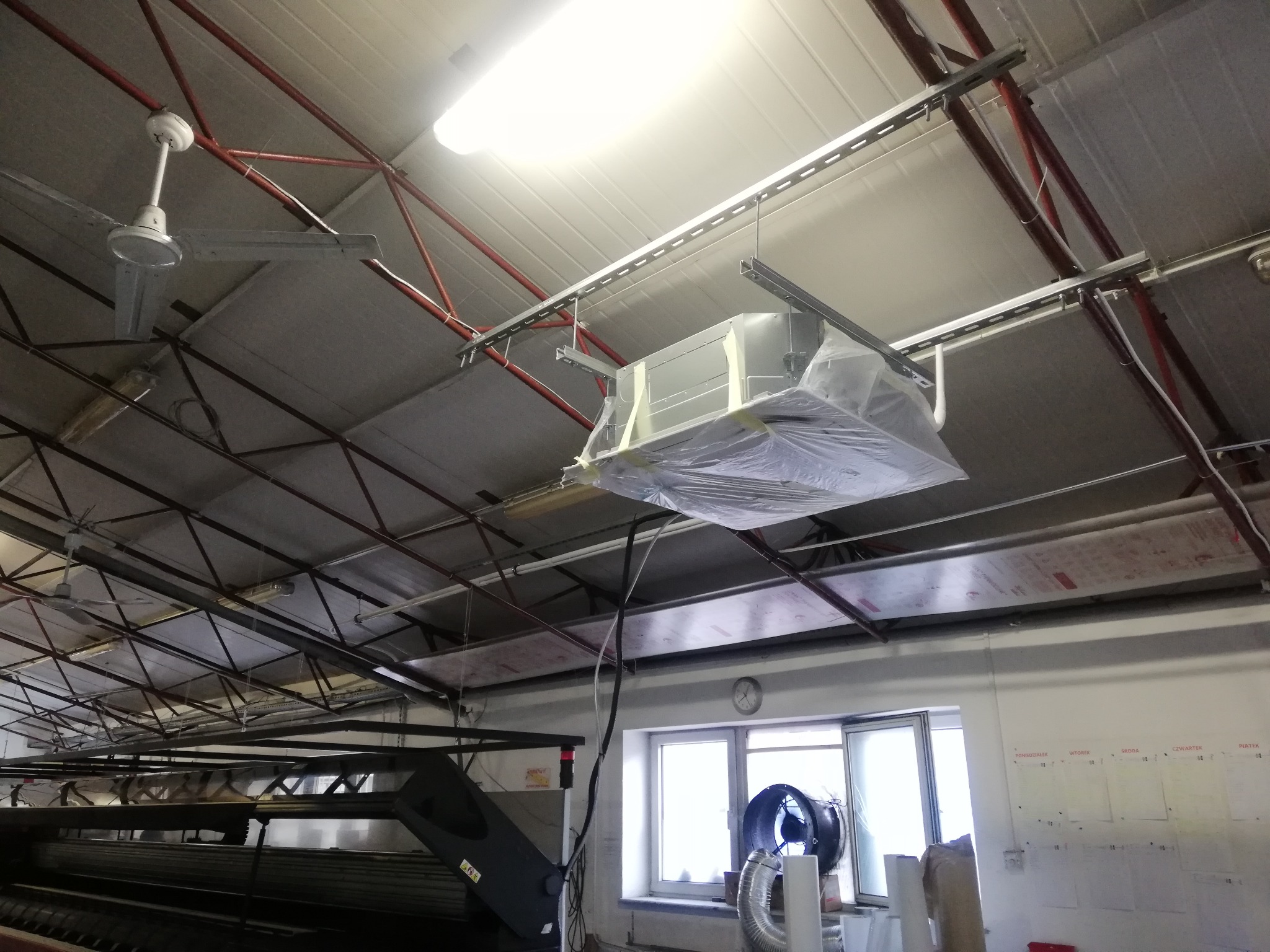 Centrala wentylacyjna w folii ochronnej, zawieszona na metalowych profilach pod sufitem hali przemysłowej. Widoczne elementy instalacji, okno z wentylatorem i kalendarz na ścianie.