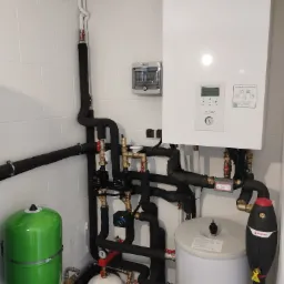 Instalacja grzewcza w kotłowni z białymi płytkami, widoczny biały zbiornik buforowy, zielone naczynie przeponowe, czarne rury z izolacją termiczną oraz biały wiszący piec Panasonic Aquarea Smart...