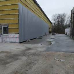 Budynek przemysłowy w trakcie ocieplania, widoczne fragmenty izolacji z wełny mineralnej i montaż blachy trapezowej na elewacji, betonowy plac przed budynkiem.