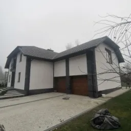 Nowoczesny dom jednorodzinny z białymi ścianami i grafitowymi akcentami z kamienia elewacyjnego, drewniane bramy garażowe, szara kostka brukowa na podjeździe, pochmurne niebo w tle.