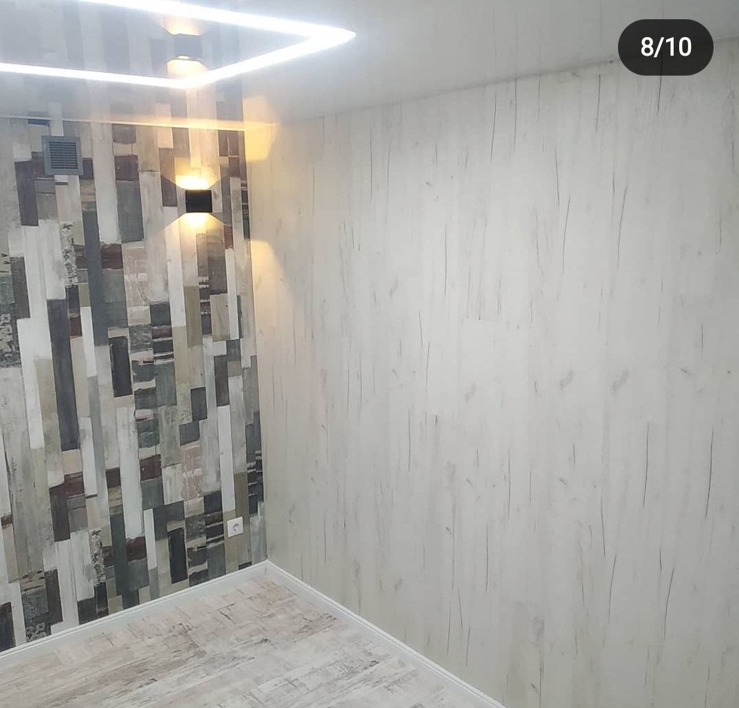 Wnętrze pokoju z dwiema ścianami o różnym wykończeniu: jedna z paneli imitujących drewno w różnych odcieniach szarości i brązu, oświetlona podwójnym kinkietem; druga pokryta tapetą w jasnym...