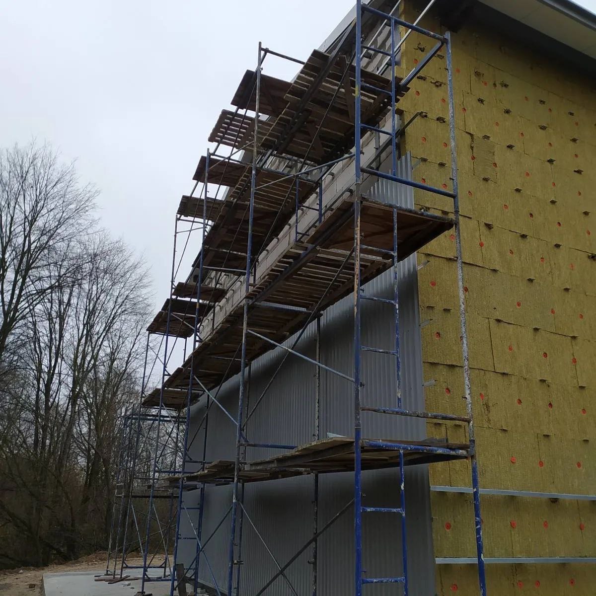 Ocieplanie ściany budynku wełną mineralną, widoczne rusztowanie i metalowe profile mocujące.