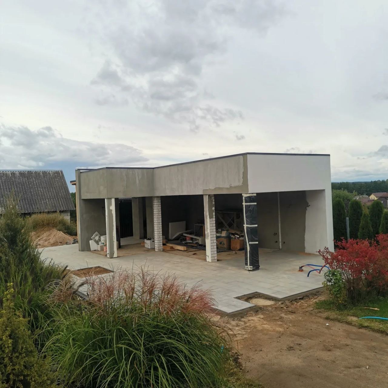 Budynek w trakcie wykończenia, widoczne szare ściany z surowym tynkiem, białe kolumny, częściowo ułożona szara kostka brukowa na podjeździe, w tle zielona roślinność i pochmurne niebo.