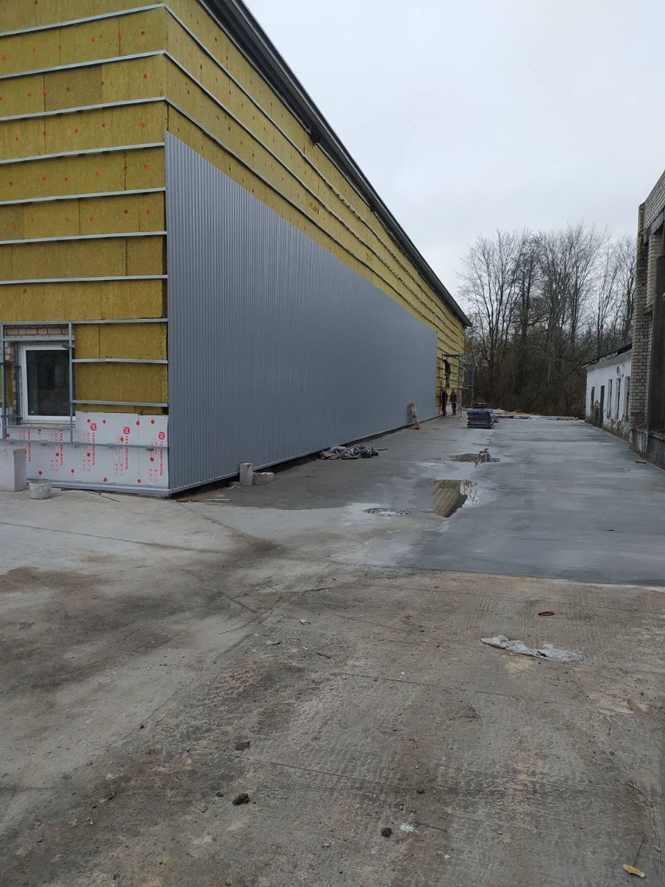 Budynek przemysłowy w trakcie ocieplania, widoczne fragmenty izolacji z wełny mineralnej i montaż blachy trapezowej na elewacji, betonowy plac przed budynkiem.