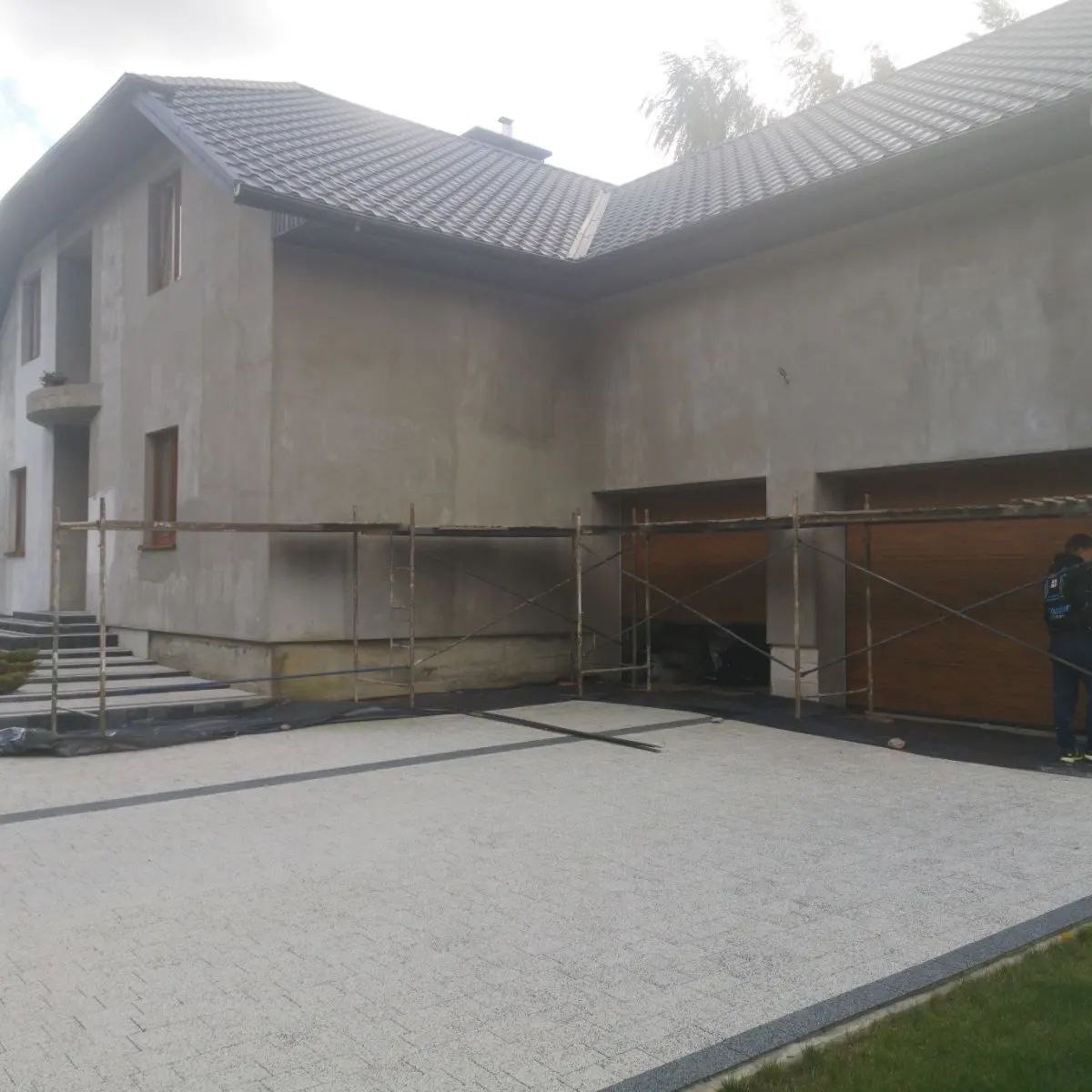 Surowa elewacja domu jednorodzinnego z dwoma bramami garażowymi w trakcie prac wykończeniowych, widoczne rusztowania i częściowo ułożona kostka brukowa na podjeździe.