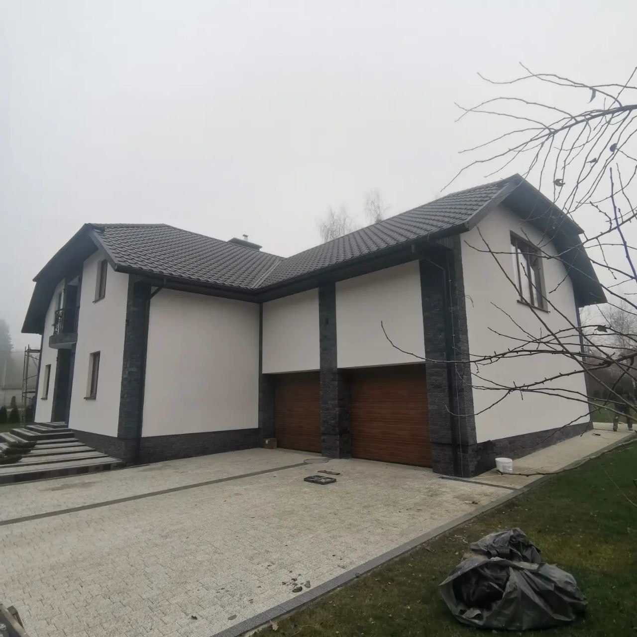 Nowoczesny dom jednorodzinny z białymi ścianami i grafitowymi akcentami z kamienia elewacyjnego, drewniane bramy garażowe, szara kostka brukowa na podjeździe, pochmurne niebo w tle.